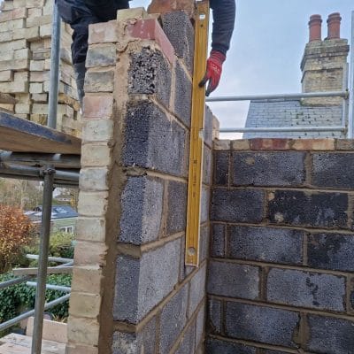 Cavity wall under construction with a dense concrete block inner leaf and a yellow gault brick outer leaf turning the corner, plumb being checked with a spirit level